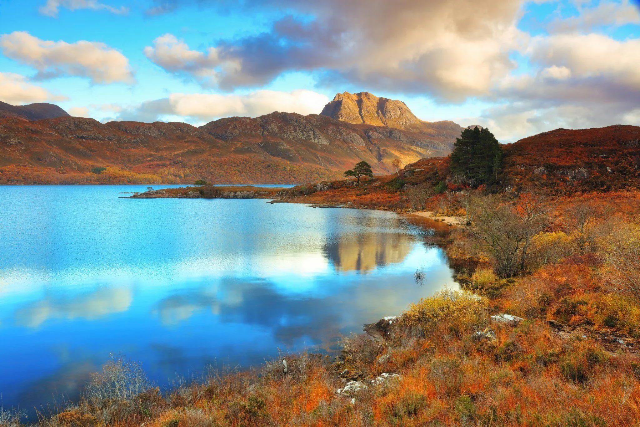 The image displays a well-composed and visually stunning natural landscape, featuring a large body of water reflecting the sky and distant mountains. The lighting is excellent, highlighting the dramatic textures of the mountains and the vibrant autumn colors of the foreground foliage. The image is sharp, and key elements are in focus, contributing to its strong photo-realistic quality. The colors are striking and appear accurate for the scene, albeit with a slight artistic saturation.