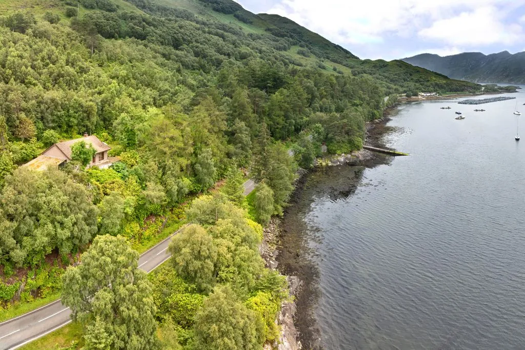 An aerial view of a scenic coastal road winding alongside a body of water, bordered by dense green forests and distant mountains under an overcast sky. The image captures the natural beauty of the landscape.