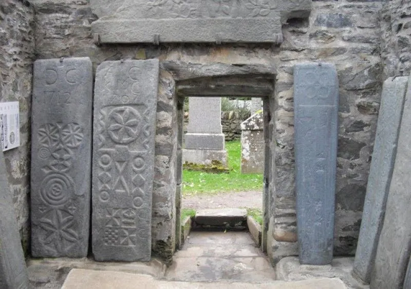 An image capturing a stone doorway within an old stone structure, flanked by two large, intricately carved standing stones. Through the doorway, a grassy area with additional stone monuments is visible. The scene suggests an ancient or historical setting, possibly a monastic site or a graveyard.
