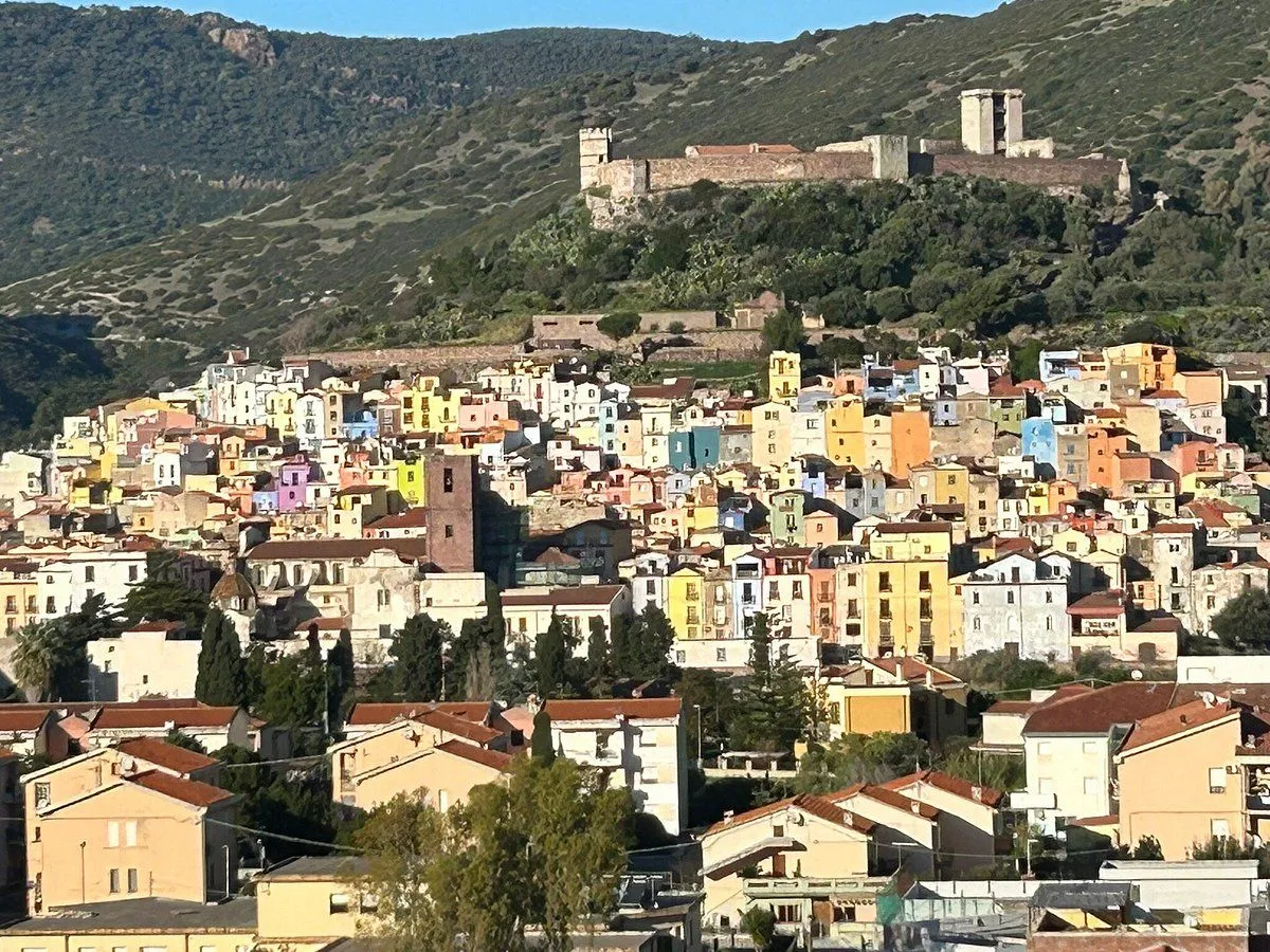 A vibrant, colorful town sprawls across a hillside, topped by a historic castle, under a clear, bright sky. The image captures the unique architectural style and dense settlement of the area.