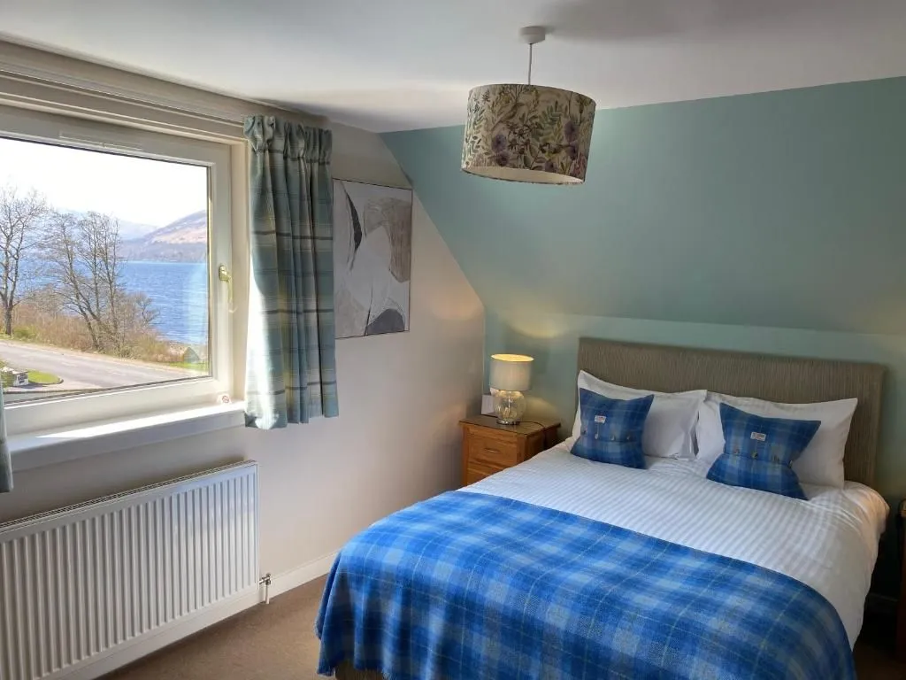 An interior shot of a well-appointed bedroom featuring a bed with blue plaid bedding, a wooden nightstand, and a prominent window offering a scenic view of a lake and distant mountains. The room is decorated with light blue and white walls, and a patterned ceiling lamp.