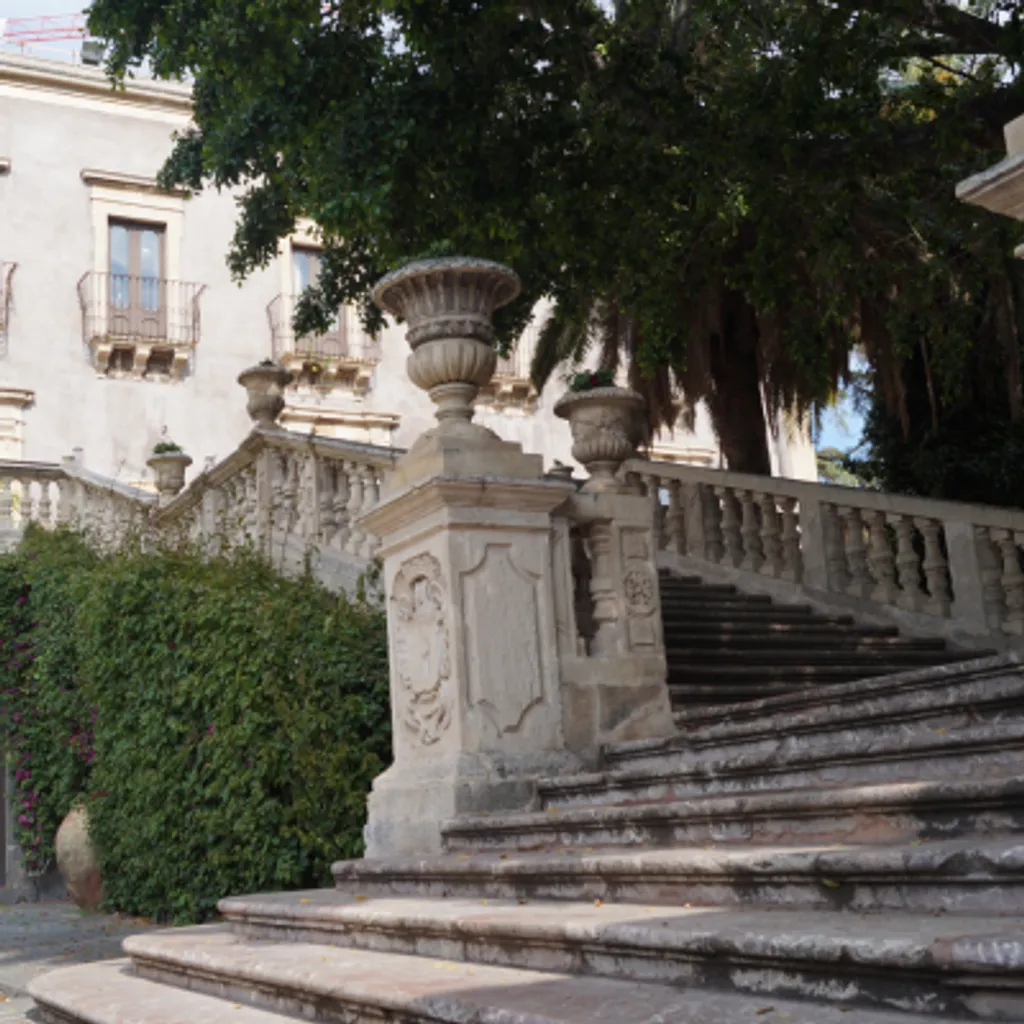 An outdoor shot of an ornate stone staircase leading up, flanked by lush green bushes and partially obscured by large trees. A historic-looking building is visible in the background.