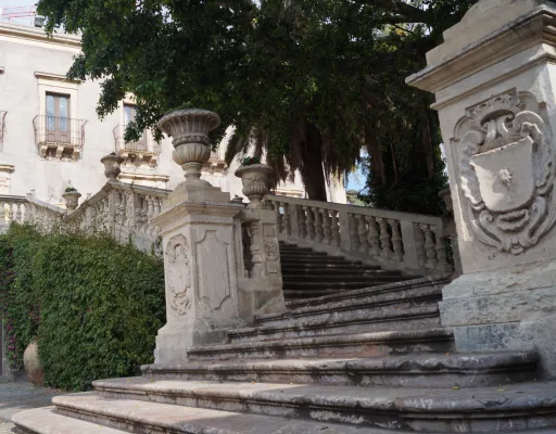 An outdoor shot of an ornate stone staircase leading up, flanked by lush green bushes and partially obscured by large trees. A historic-looking building is visible in the background.