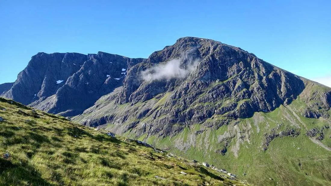 A vibrant and clear landscape image featuring a large, rugged mountain range under a bright blue sky. The foreground shows grassy slopes, leading up to the rocky mountains with patches of green vegetation and some remnants of snow. A small cloud hovers near the peak of the central mountain, adding to the scenic view.