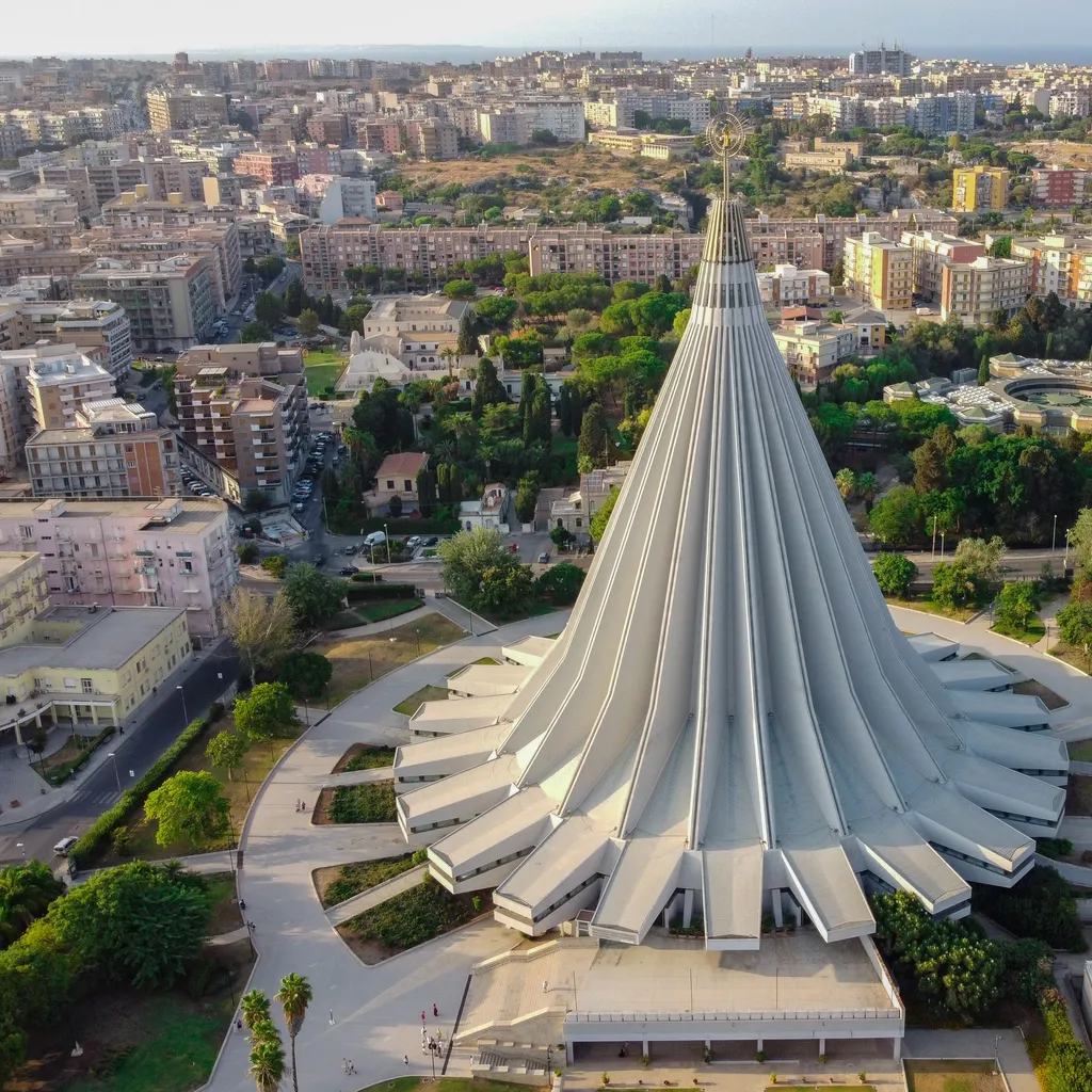 An excellent aerial shot showcasing the unique, modern architecture of the Sanctuary of Our Lady of Tears in Syracuse. The composition is strong, centering the distinctive conical structure against a backdrop of the city.