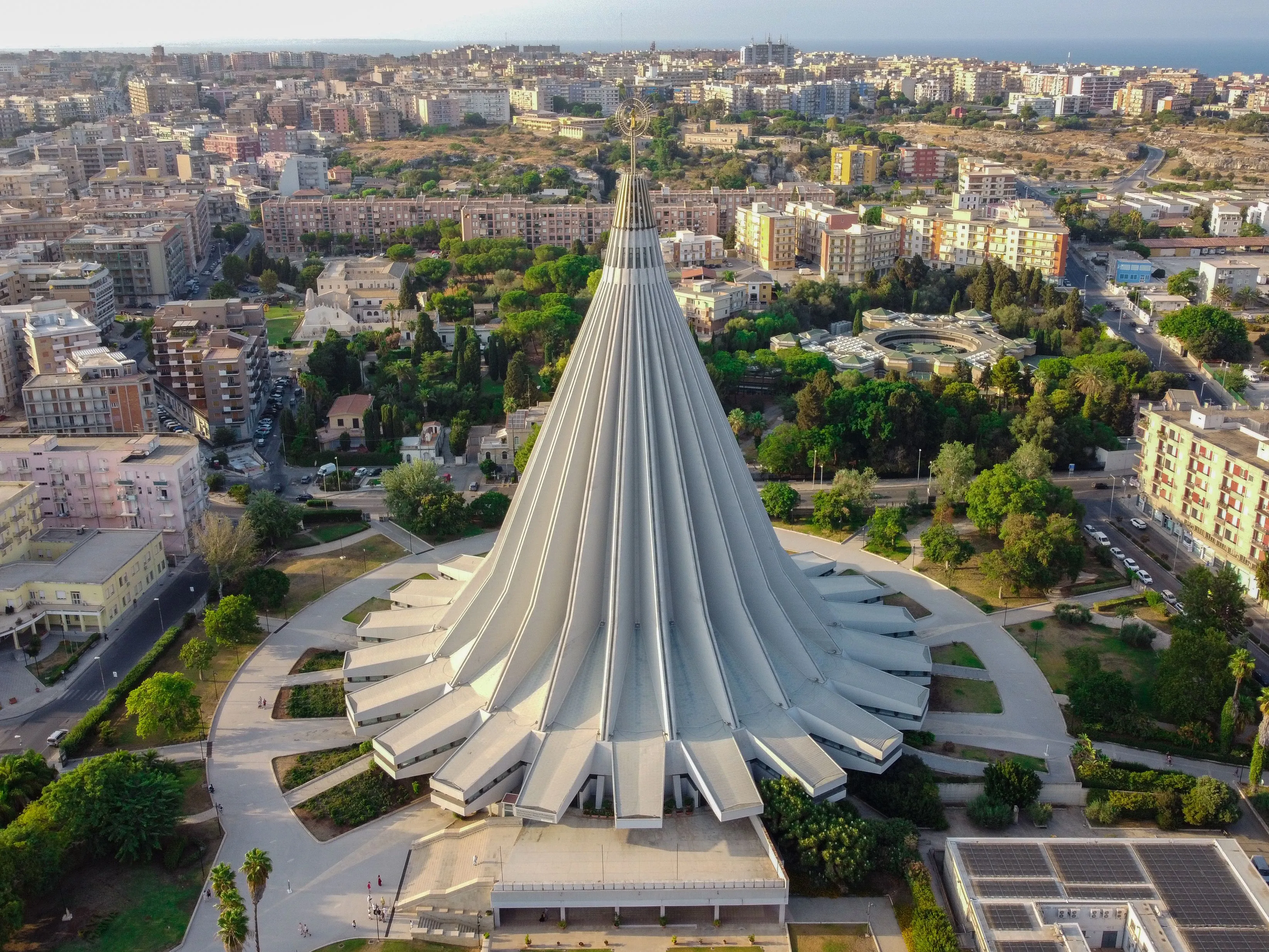 An excellent aerial shot showcasing the unique, modern architecture of the Sanctuary of Our Lady of Tears in Syracuse. The composition is strong, centering the distinctive conical structure against a backdrop of the city.