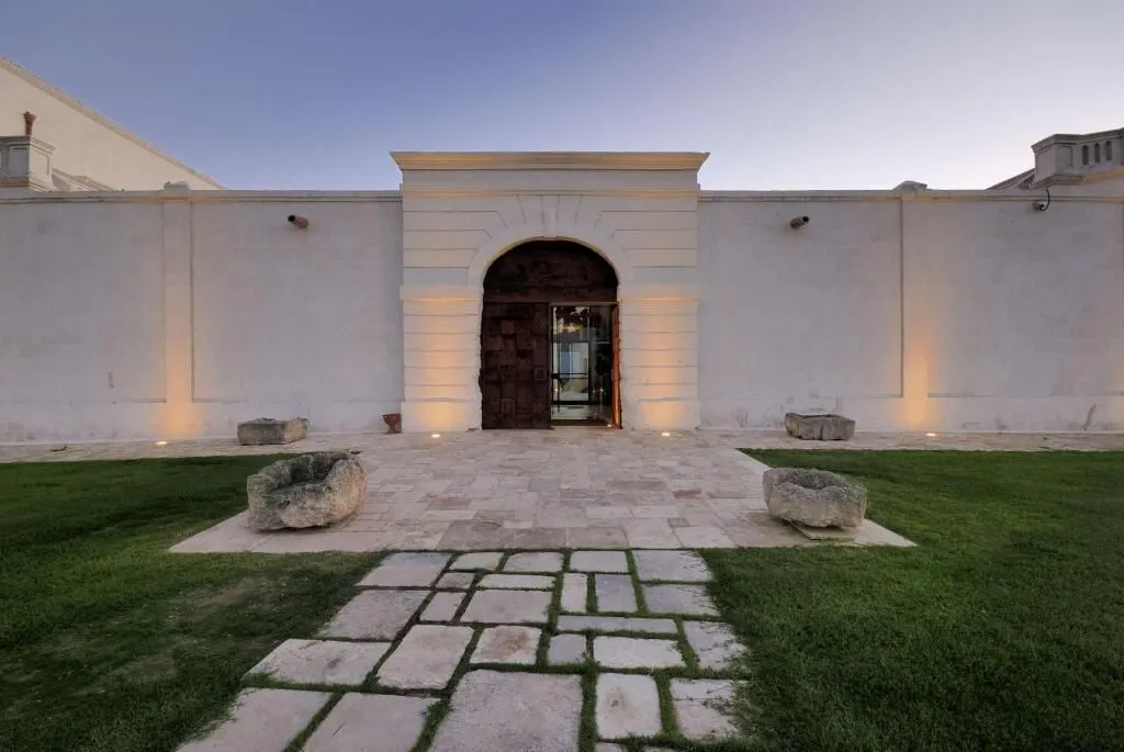 An exterior view of a building at dusk or dawn, featuring a large arched entrance, a stone pathway, and well-maintained grassy areas. Ground-level lights illuminate the facade and entrance.