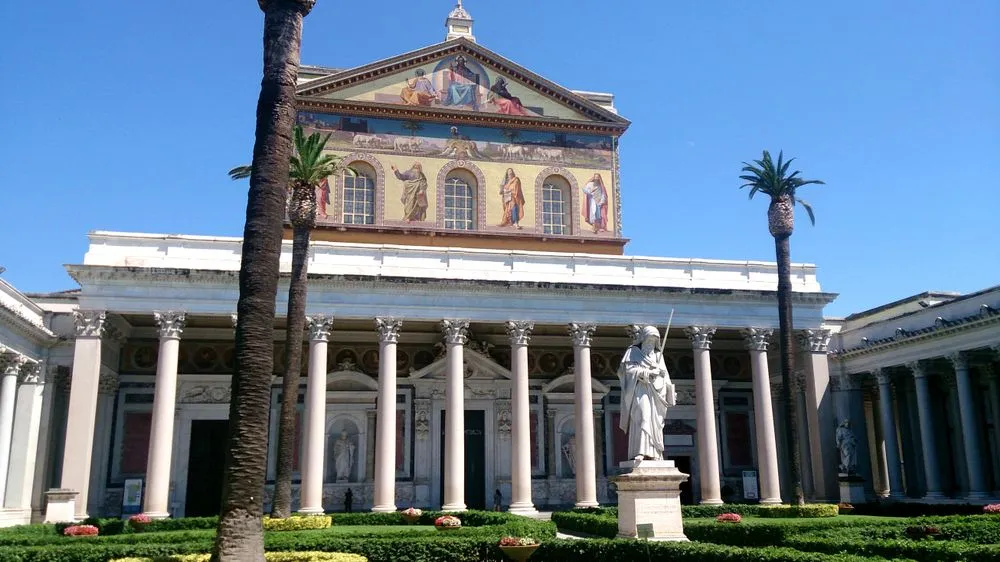The image captures a grand architectural structure, likely a basilica or church, with classical columns and intricate facade details, set against a clear blue sky. A statue stands in the foreground, with well-maintained greenery and palm trees surrounding the building.