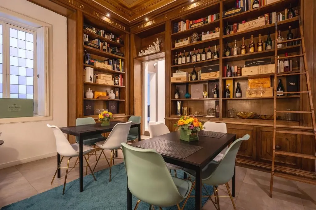 An inviting interior view of a room featuring extensive wooden bookshelves filled with books and wine bottles, along with modern dining tables and chairs.