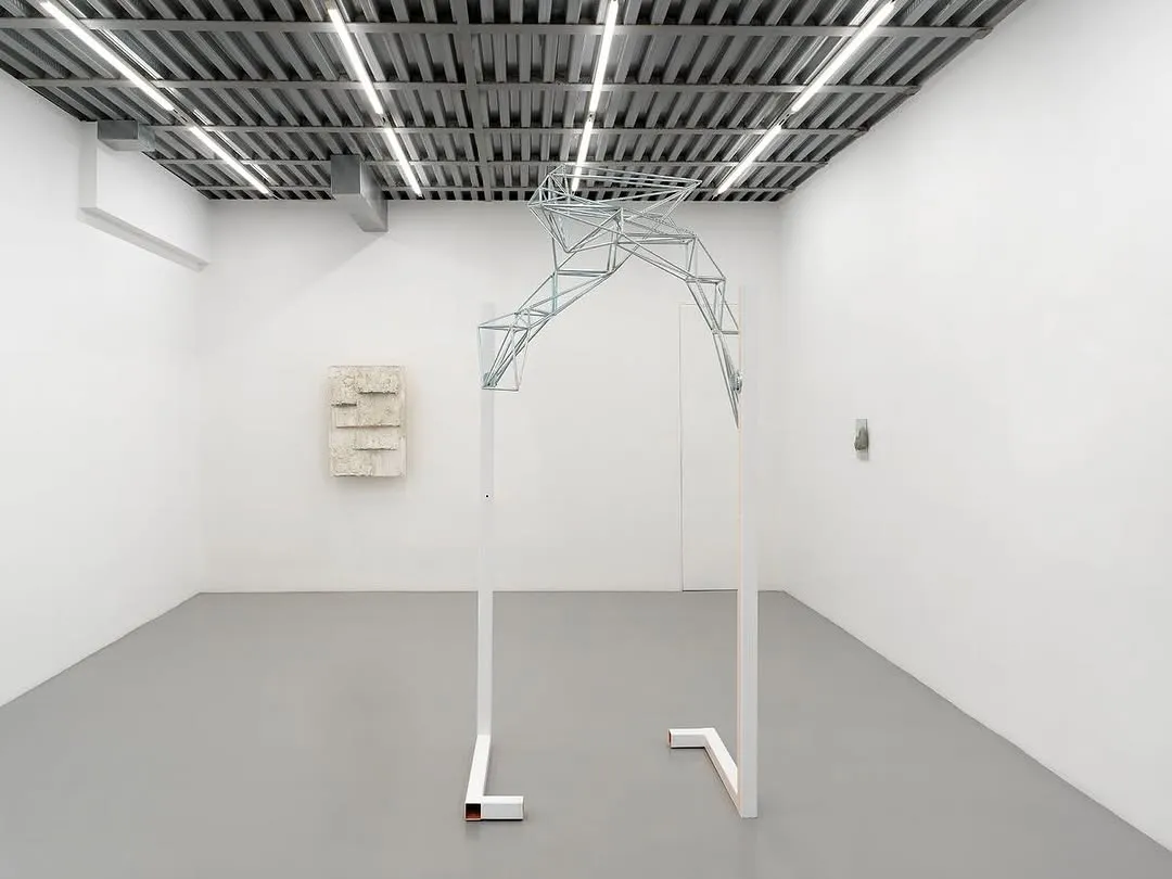 A photograph of a contemporary art installation in a minimalist gallery space, featuring a large, abstract metal sculpture resembling an arch or skeletal structure, with other smaller artworks visible on the white walls.