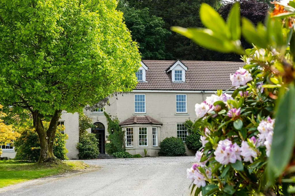 An outdoor view of a classic two-story house with a bay window and dormer windows, featuring a tiled roof and light-colored facade, nestled among lush green trees and flowering shrubs. A gravel driveway leads to the front entrance.