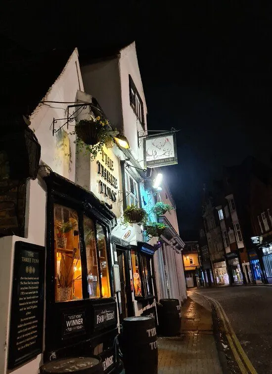 A nighttime shot of 'The Three Tuns' pub exterior, highlighting its illuminated facade and signboard against a dark street and sky. The image captures the atmospheric glow of the pub lights.