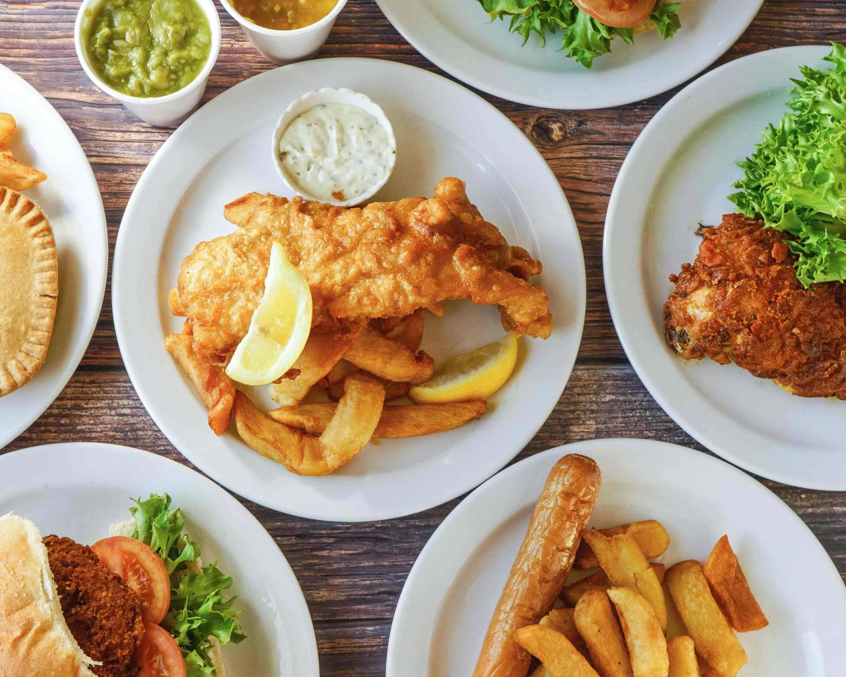 The image is a well-composed overhead shot featuring a variety of fried foods, with a plate of fish and chips prominently in the center. The lighting is excellent, ensuring all elements are clearly visible and appetizing. The image is sharp with accurate, vibrant colors, contributing to a highly photo-realistic appearance. Key elements, particularly the fish and chips, are perfectly in focus. The overall presentation is professional and visually appealing, fitting an editorial style.