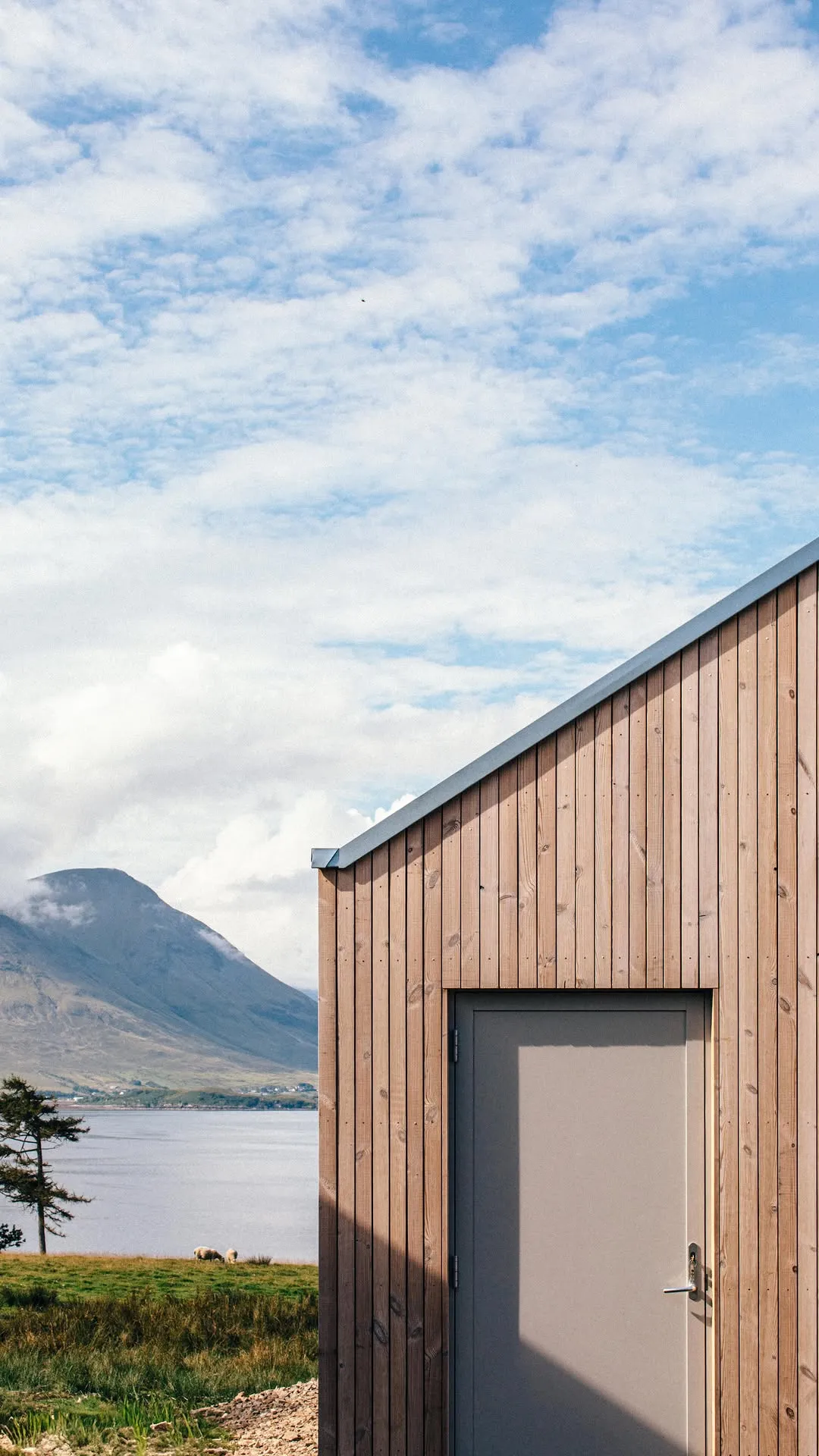 A modern building with wooden cladding and a gray roof stands prominently in the foreground, with a large mountain and a body of water visible in the background under a partly cloudy sky.