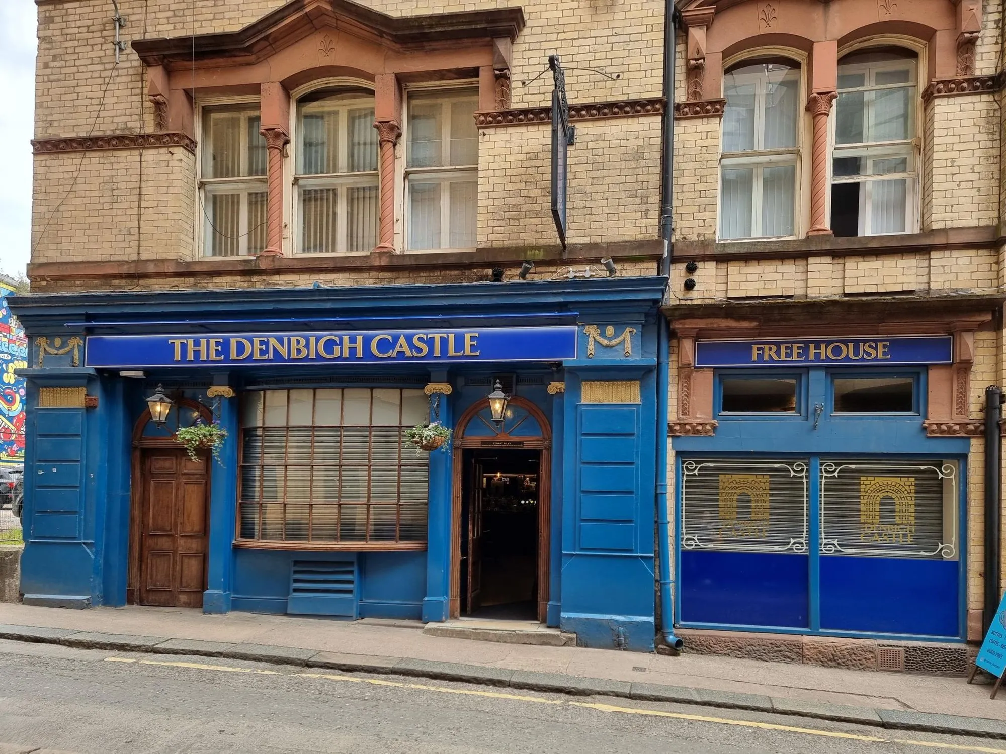 An image of the exterior of 'The Denbigh Castle' pub, featuring its distinctive blue facade, windows, and signage. The building is made of brick with ornate architectural details.