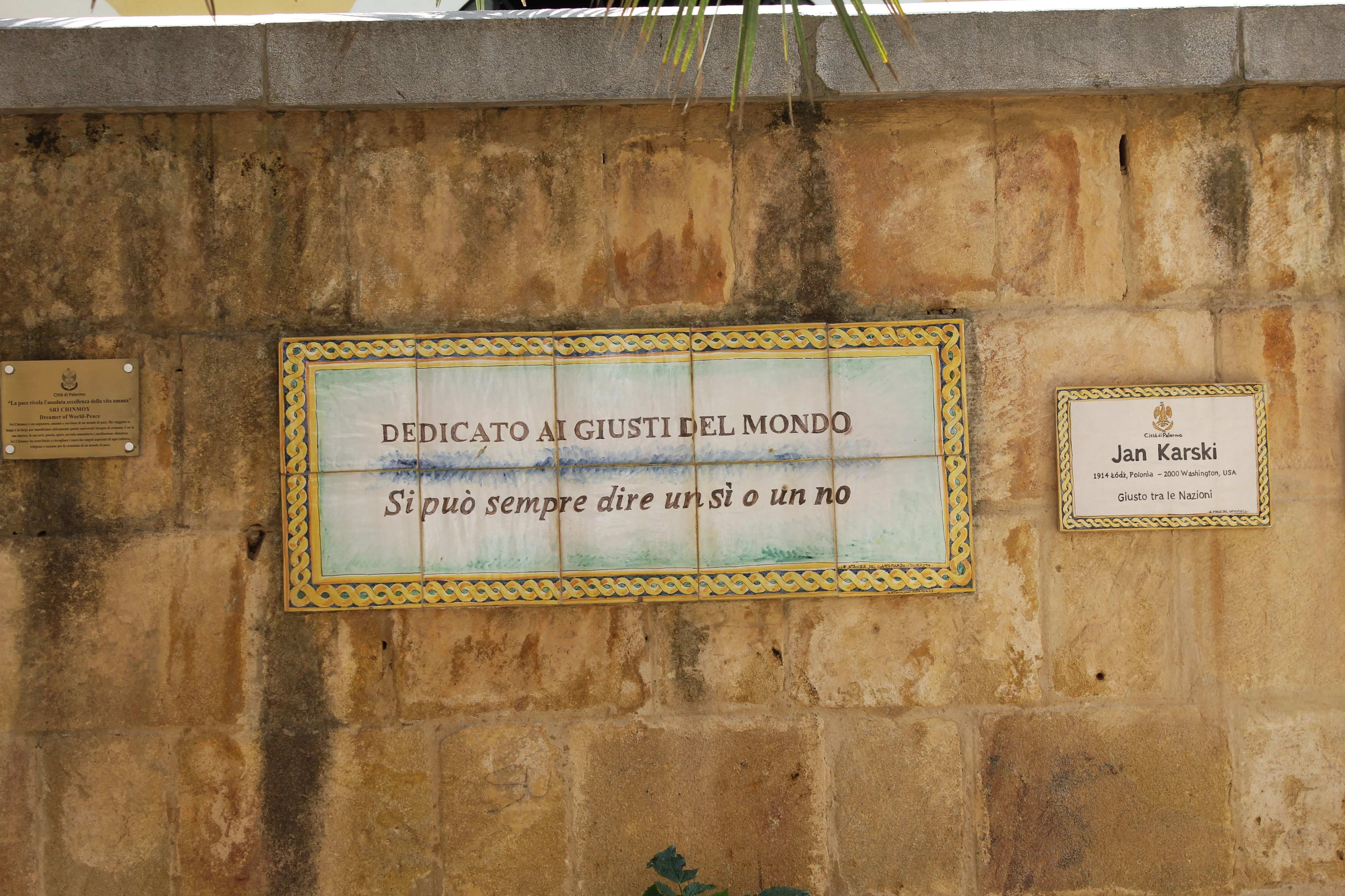The image features a ceramic plaque mounted on a stone wall, likely a dedication. The lighting is natural and even, contributing to good color accuracy. The plaque and its text are sharp and clearly in focus, enhancing photo-realism. Composition is generally good, with the main subject centered, although it could be slightly improved by ensuring the plaque is perfectly straight and avoiding the slight crop at the top and bottom, as well as the partial visibility of another plaque on the right side.