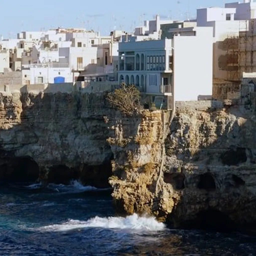 The image is a well-composed and vibrant depiction of a coastal town situated atop rugged cliffs overlooking the sea. The natural lighting is excellent, illuminating the scene effectively and enhancing the textures of the buildings and cliffs. The image is sharp with clear details, and the colors, particularly the blues of the sea and the whites of the buildings, appear accurate and natural. The photograph is highly realistic, and all key elements are in sharp focus.