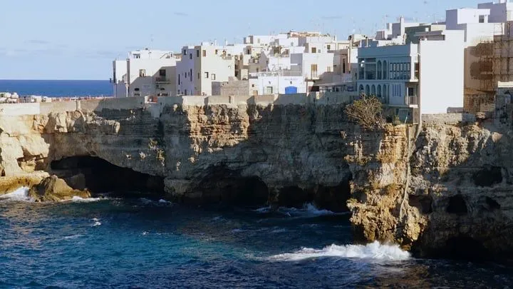 The image is a well-composed and vibrant depiction of a coastal town situated atop rugged cliffs overlooking the sea. The natural lighting is excellent, illuminating the scene effectively and enhancing the textures of the buildings and cliffs. The image is sharp with clear details, and the colors, particularly the blues of the sea and the whites of the buildings, appear accurate and natural. The photograph is highly realistic, and all key elements are in sharp focus.