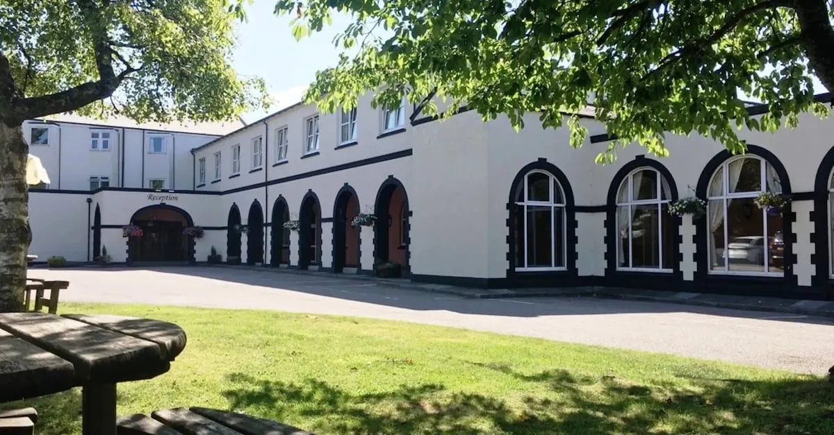 An exterior shot of a building featuring multiple arched entrances and windows, set against a backdrop of green grass and trees under clear skies. The building is light-colored with dark trim around the arches and windows.