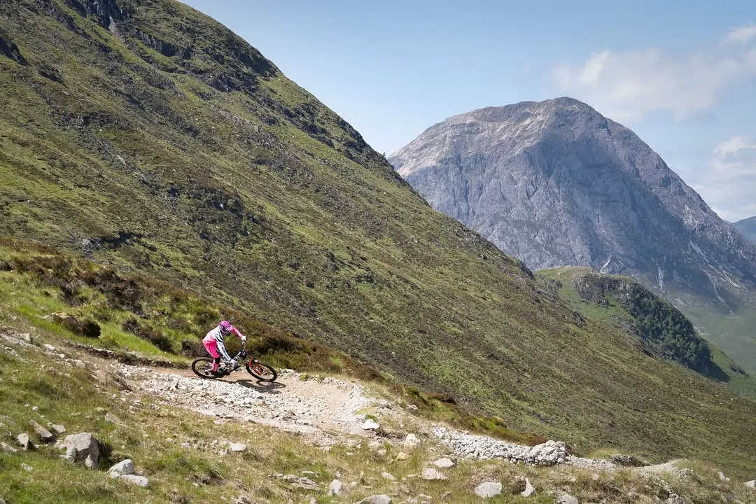 An action shot of a mountain biker on a rocky trail with a vast mountain landscape in the background. The image is well-composed with good leading lines, and the lighting is natural and even. The subject is sharp and in focus, contributing to the photo's realism and clarity.