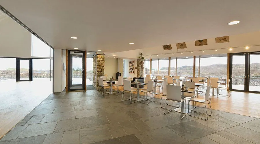 An interior shot of a modern cafe or restaurant space, featuring large windows that offer natural light and a view of the outdoors. The room is furnished with multiple tables and chairs, and includes architectural details like stone pillars.