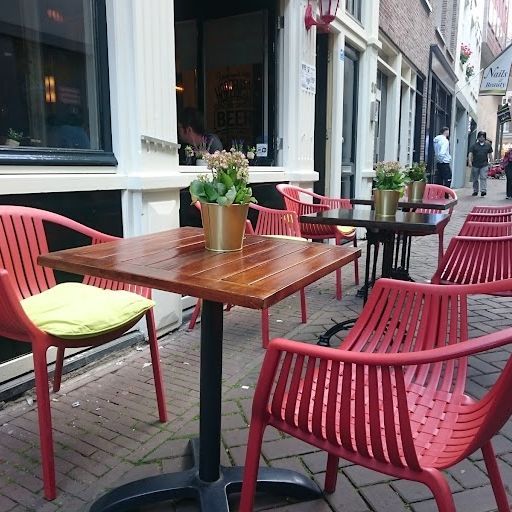 The image is an example of exterior photography. It shows a terrace with tables, chairs, and flowers. The composition is simple and balanced, with the main focus on the red chair in the foreground. The image is well-lit and appears to be taken during the day. The colors are accurate, and the image is sharp. There are some unwanted elements in the image, such as the people walking in the background and the sign on the door. However, these elements do not detract from the overall quality of the image.