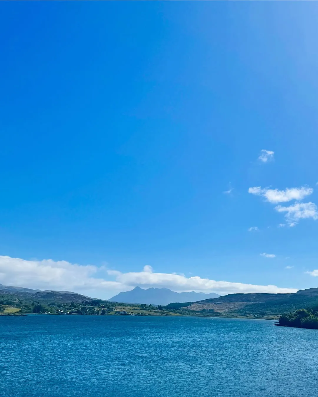 The image is a clear and vibrant landscape shot featuring a vast expanse of blue water, distant mountains, and a bright blue sky with sparse clouds.