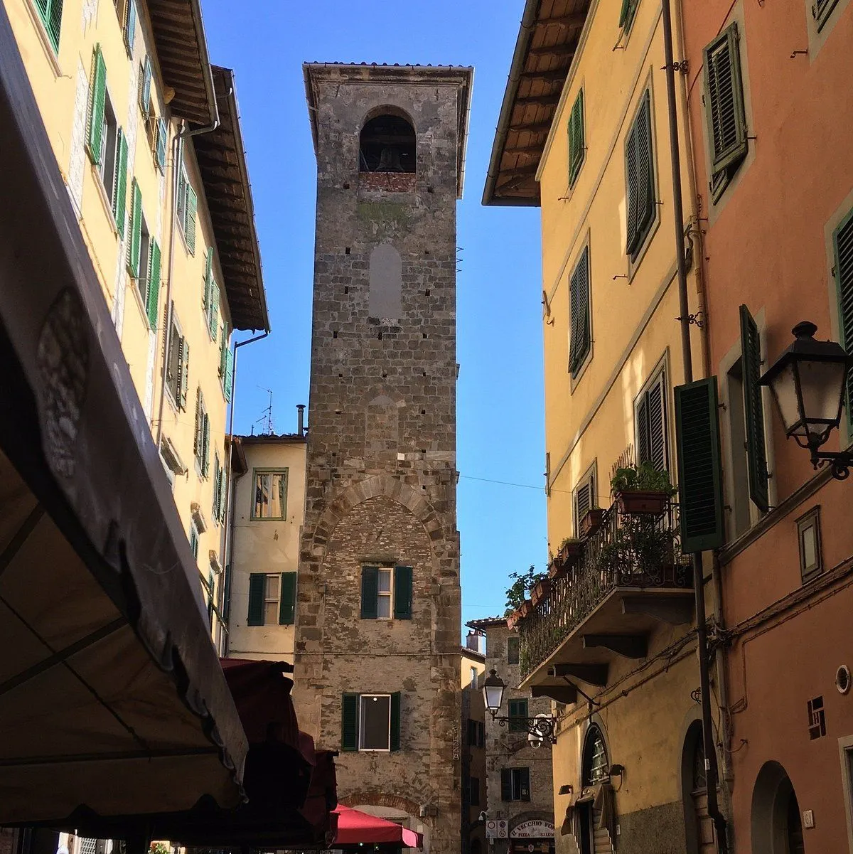 A tall, historic stone tower stands prominently at the end of a narrow street, framed by traditional European buildings with colorful facades and green shutters under a clear blue sky.