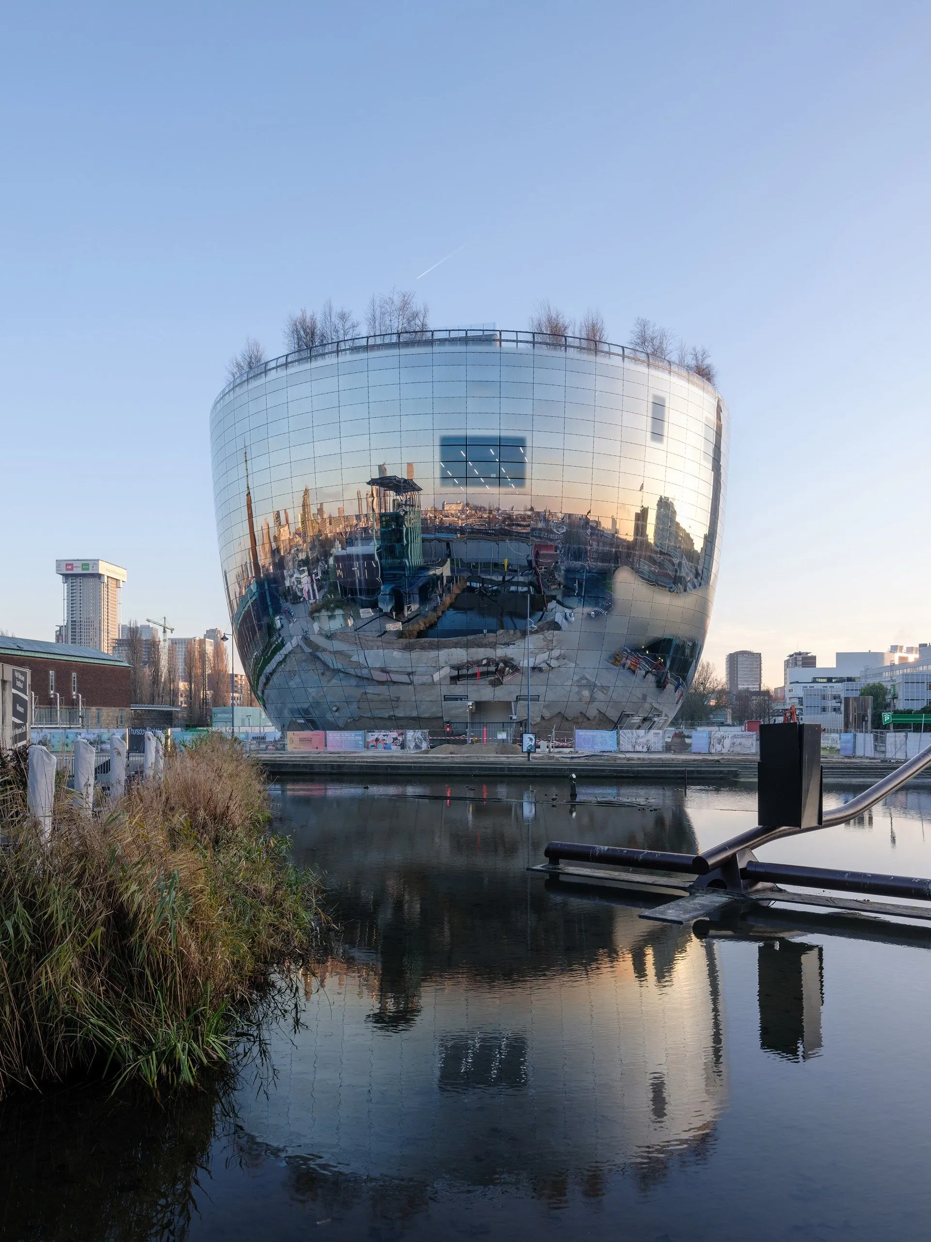 An editorial image featuring a modern, reflective building with a unique, rounded design situated next to a body of water. The building's glass facade reflects the surrounding urban landscape and the clear blue sky, creating an intriguing visual effect. Reeds are visible in the foreground, and distant city structures can be seen under a bright, natural light.