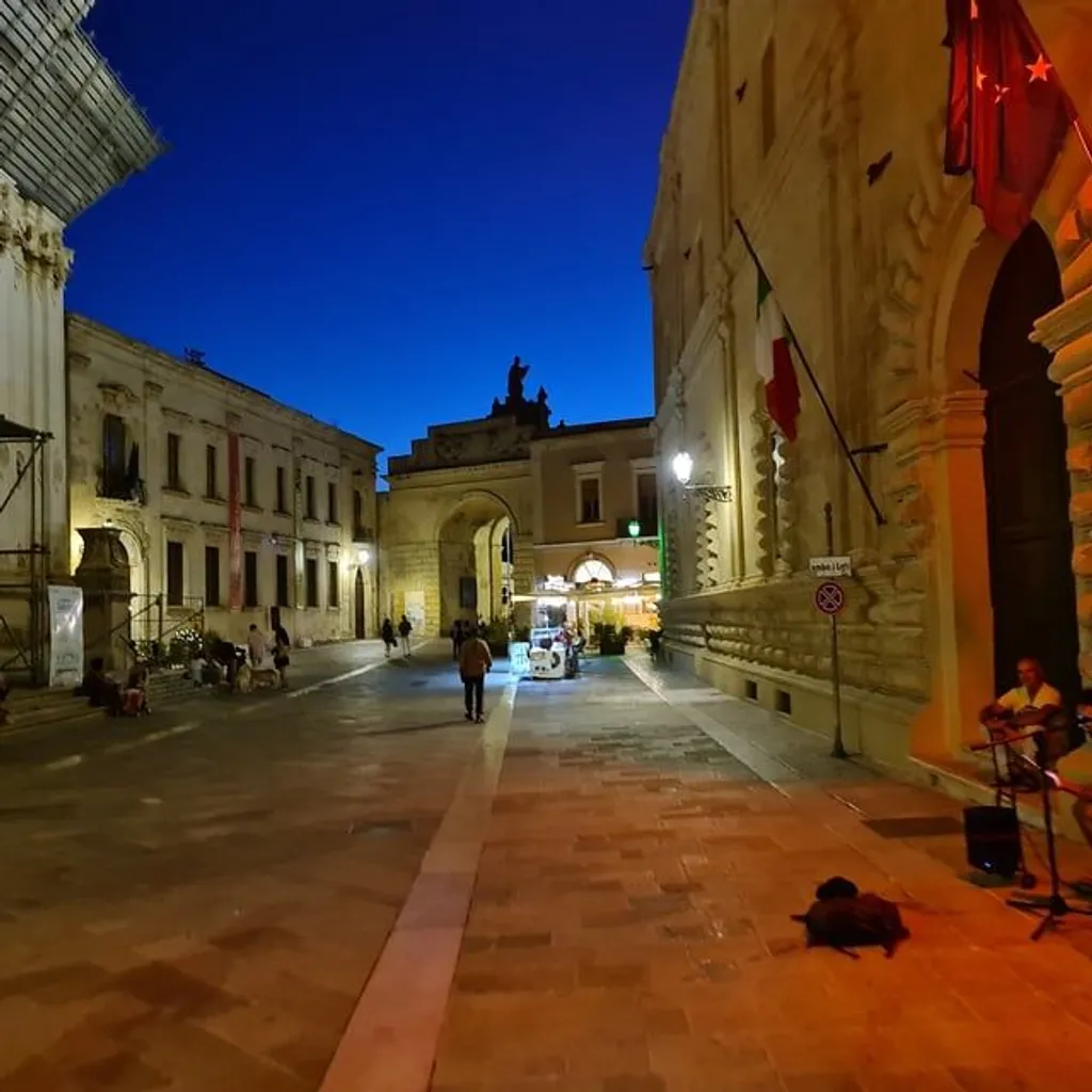 A night-time view of an old town street, featuring historic buildings illuminated by artificial lights under a deep blue twilight sky. The street, paved with light-colored stones, leads towards an archway in the distance. People are present on the street, with a street musician visible on the right.
