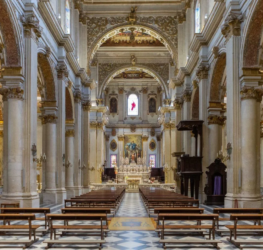 An interior shot of a grand and ornate church or cathedral, showcasing the long nave leading towards the altar, flanked by rows of pews and intricate architectural details including columns, arches, and decorative ceilings.