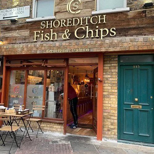 An exterior shot of 'Shoreditch Fish & Chips,' showcasing the storefront, signage, and an outdoor seating area with a person partially visible inside.
