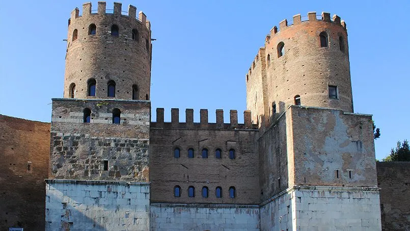 An editorial-style image featuring an old castle structure with two prominent towers and a connecting wall against a clear blue sky. The image focuses on the architectural details of the brickwork and stone.