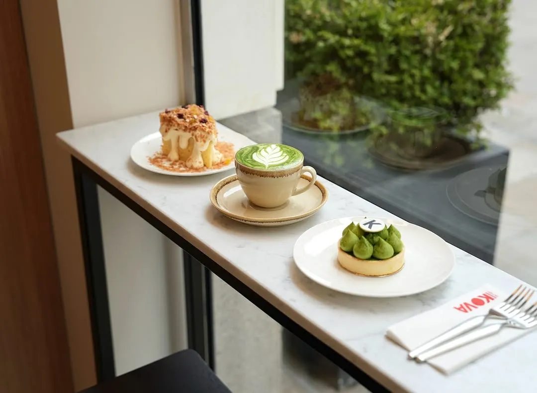 A well-composed shot featuring a matcha latte, a layered cake with crumble, and a green tart arranged on a white marble counter next to a window, illuminated by natural light.