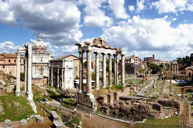 An editorial-style photograph capturing a panoramic view of the ancient Roman Forum in Rome, Italy, under a partly cloudy sky. The image features prominent ruins, including the columns of the Temple of Saturn and other historical structures, with a path winding through the archaeological site.