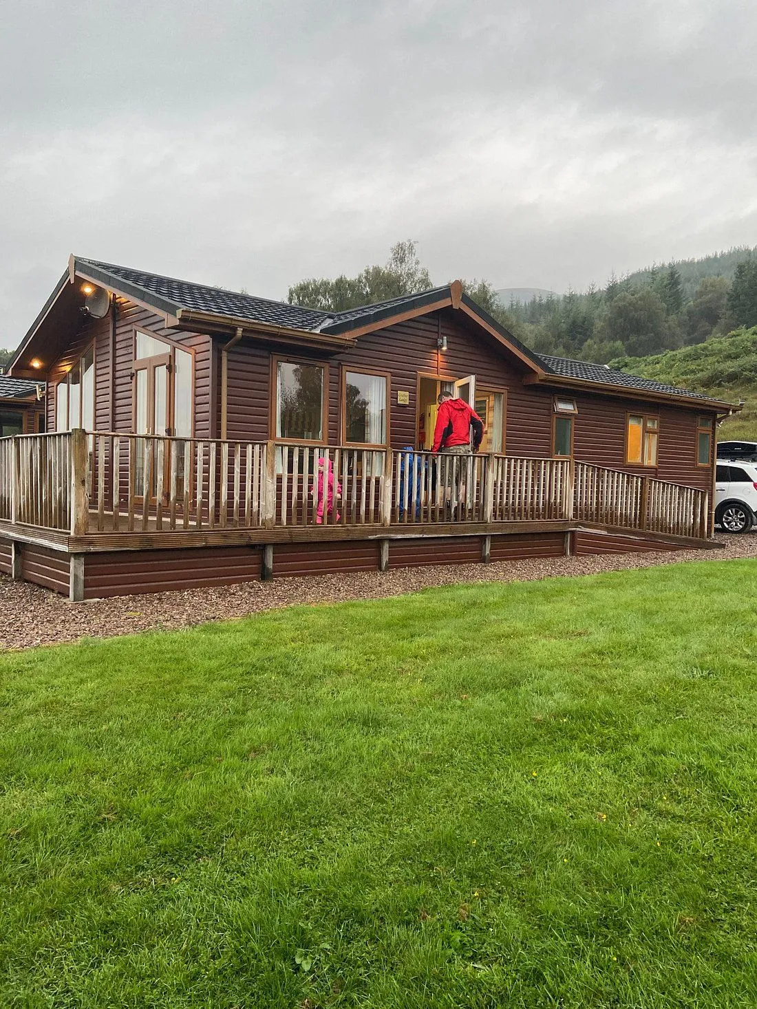 An image featuring a brown wooden holiday lodge with a large deck and green lawn in the foreground, set against a backdrop of trees and hills under an overcast sky.