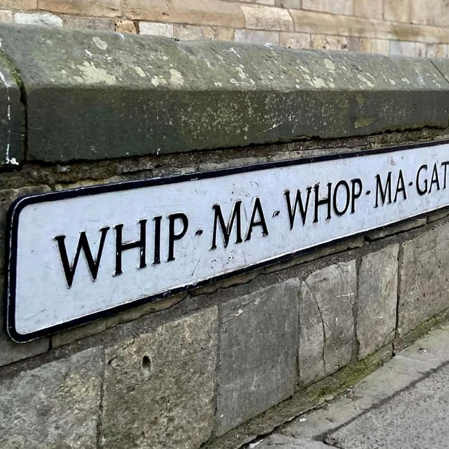The image clearly depicts the 'Whip-Ma-Whop-Ma-Gate' street sign, which is the primary subject and is in sharp focus. The composition is effective, drawing attention directly to the sign against a textured stone wall. Lighting is natural and even, contributing to the photo-realistic quality. Colors appear accurate. Some slight softness is noticeable in the background stone textures, but the key element remains highly defined.