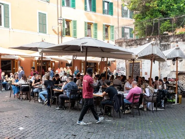 The image captures a vibrant outdoor dining scene with numerous people seated at tables under large umbrellas. The setting appears to be a European city street, identifiable by the cobblestone ground and the architecture of the building in the background. The scene is bustling with activity, featuring a person in a red shirt walking through the foreground. The lighting is natural daylight, contributing to a realistic portrayal of the scene. The overall color palette is natural, although slightly muted. Key elements like the people and the dining area are generally in focus.
