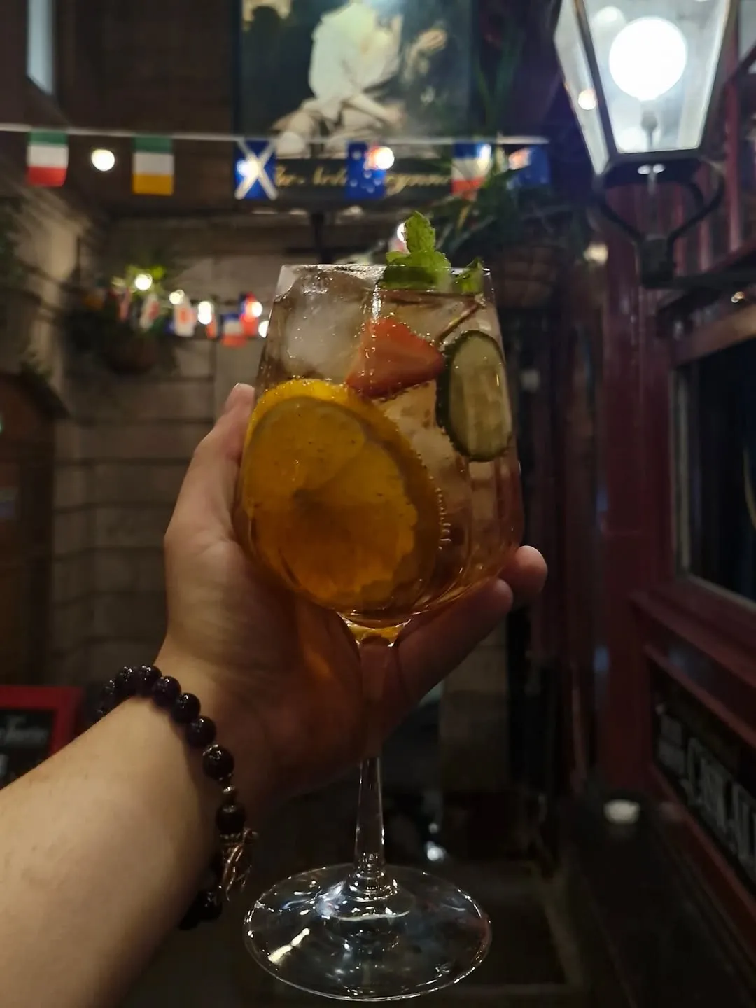 A close-up, well-lit image of a refreshing drink, likely a cocktail or spritzer, held in a hand. The glass contains ice, an orange slice, strawberry, cucumber, and mint, all appearing sharp and color-accurate. The background, though somewhat busy with flags and lights from what appears to be a pub or bar setting, adds an atmospheric touch without fully detracting from the main subject.