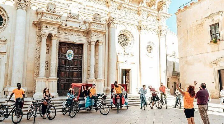The image features a group of people on bikes and pedicabs in front of an ornate, white baroque-style church building. The scene suggests a travel or tourism context, possibly a guided tour or exploration of a historic city. The lighting is bright and natural, indicating a sunny day.