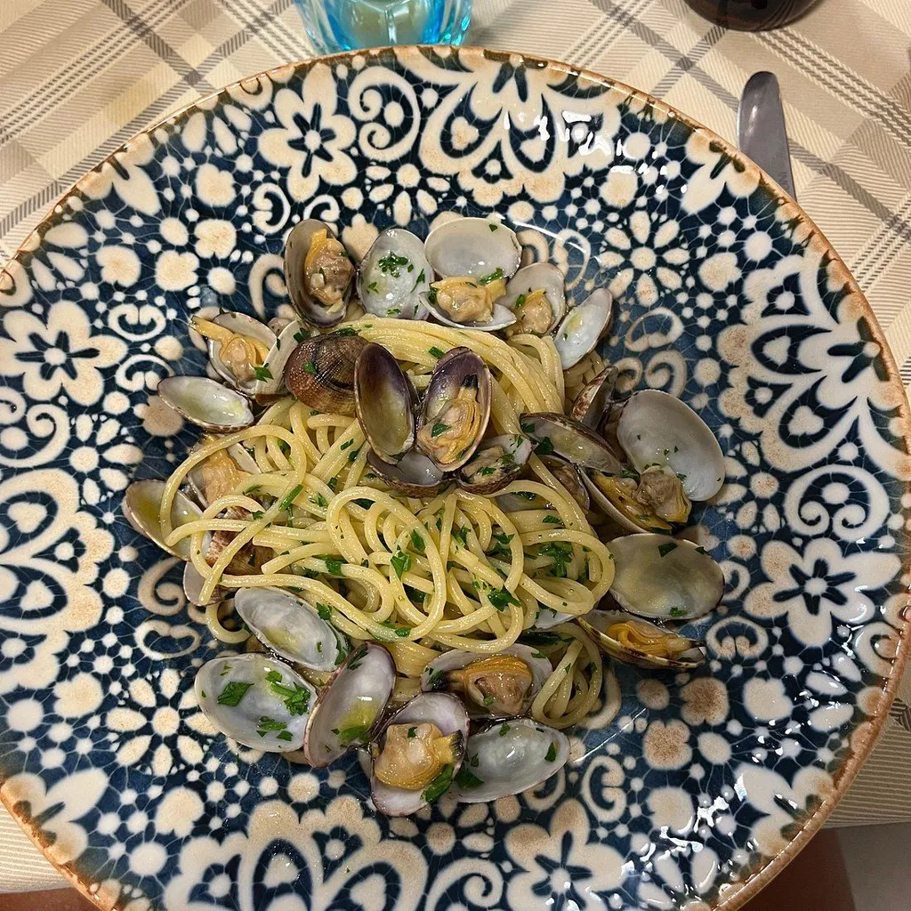 A close-up, overhead shot of spaghetti with clams served on a decorative blue and white plate. The pasta is topped with chopped green herbs. A small portion of a glass and a knife are visible in the background.
