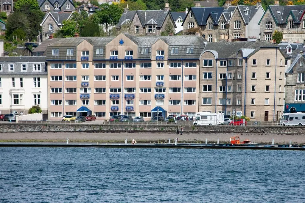 A daylight shot of a multi-story building complex situated along a waterfront, with cars parked at the street level and a body of water in the foreground. Additional buildings are visible on a hillside in the background.
