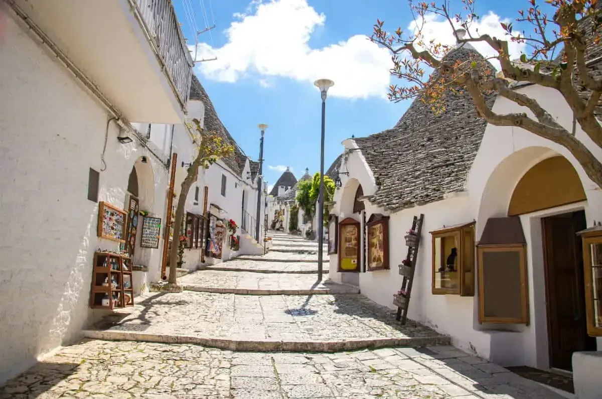 An editorial-style image of a charming, narrow street lined with traditional white-washed houses featuring unique cone-shaped stone roofs, characteristic of Alberobello, Italy. The street is paved with stones and slopes upwards, leading the eye through the scene. The sky is bright blue with some clouds.