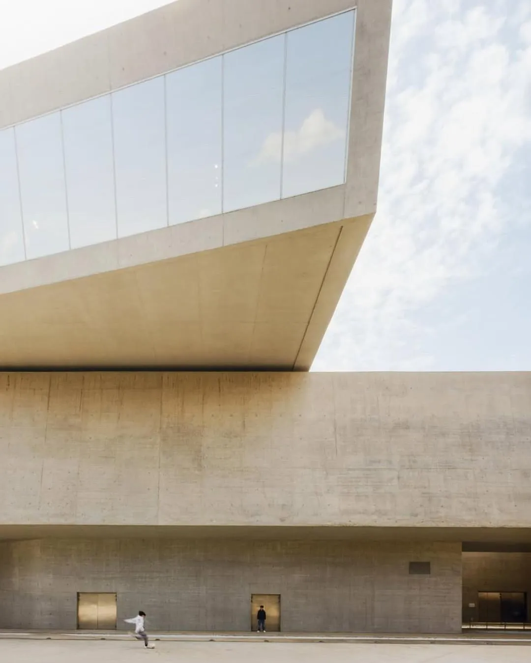 The image showcases a modern architectural structure with strong geometric lines, predominantly featuring concrete and large glass windows against a clear sky. The composition is well-balanced, emphasizing the building's form and textures. Lighting is even and natural, contributing to the overall clarity.