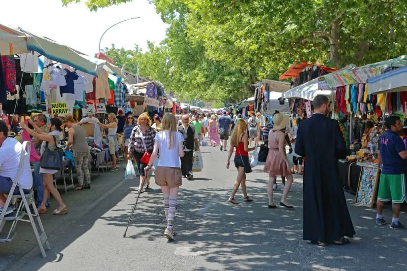 A bustling outdoor market scene with numerous people walking and browsing stalls under a canopy of trees.
