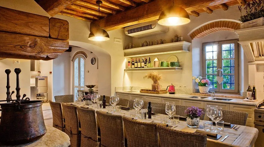 An inviting dining room setup featuring a long table with wicker chairs, set for a meal. The room is decorated with a rustic, warm aesthetic, including wooden beams on the ceiling and a well-stocked kitchen/bar area in the background with exposed brick accents and shelves displaying bottles and dishware. Warm lighting emanates from pendant lamps and under-shelf strips.