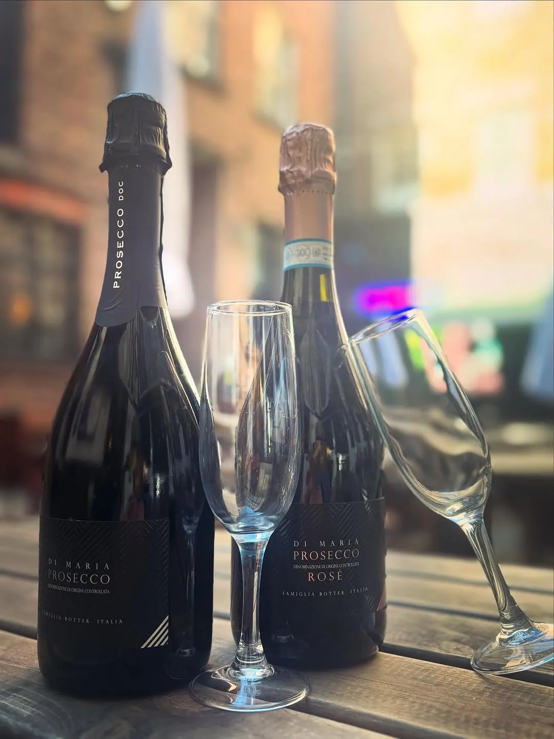 Two bottles of prosecco, one white and one rosé, are prominently displayed with two empty champagne flutes on a wooden surface. The background is softly blurred, hinting at an urban or outdoor setting.