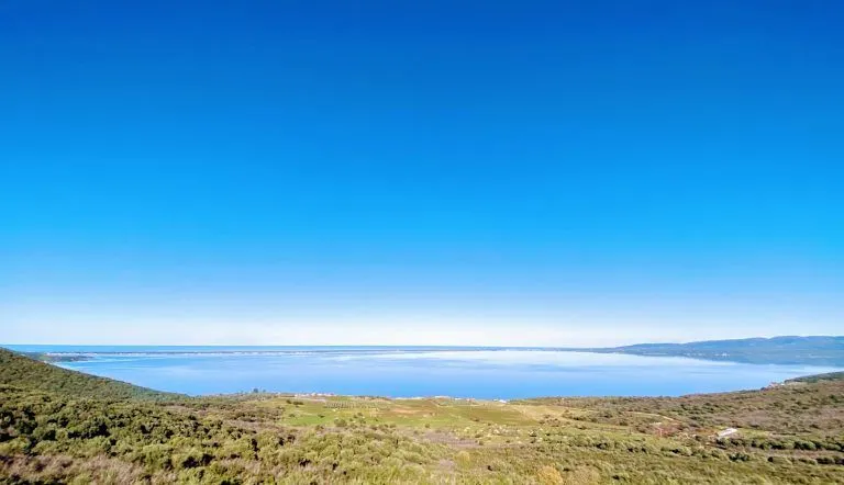 A pristine landscape image featuring a vibrant blue sky, calm blue water, and a lush green and brown foreground. The composition is simple and effective, highlighting the natural beauty of the scene.