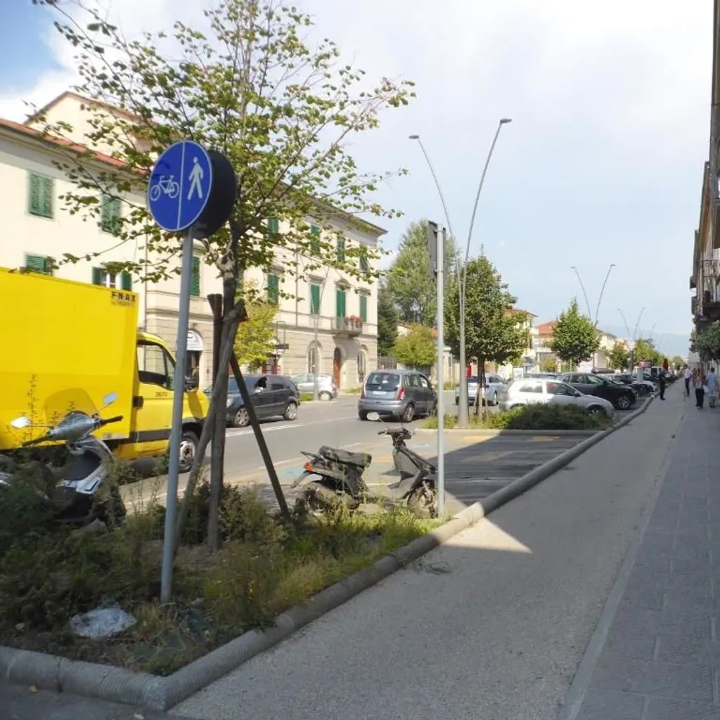 An outdoor daytime photograph capturing a street scene lined with buildings, parked cars, trees, and a sidewalk. A blue sign indicating a shared pedestrian and bicycle path is prominent in the foreground, with people visible in the distance along the sidewalk.