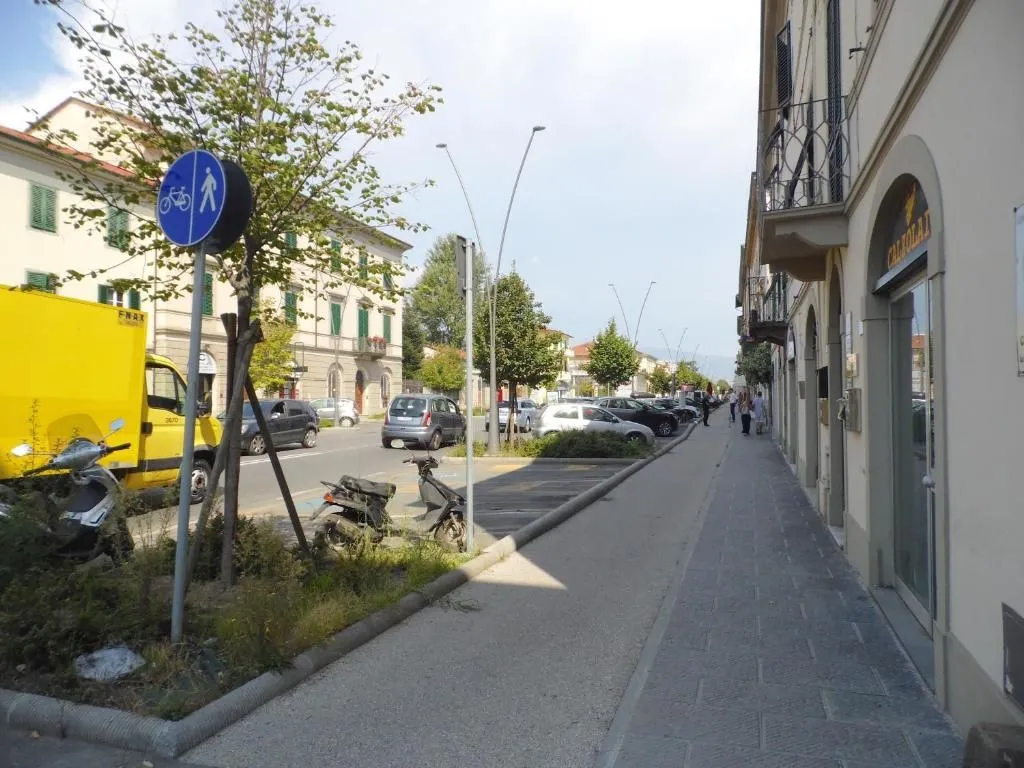 An outdoor daytime photograph capturing a street scene lined with buildings, parked cars, trees, and a sidewalk. A blue sign indicating a shared pedestrian and bicycle path is prominent in the foreground, with people visible in the distance along the sidewalk.